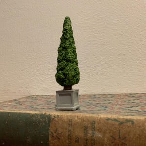Pode incluir: Uma pequena árvore topiária verde num vaso quadrado cinzento. A árvore está colocada num livro vintage com uma capa estampada. A árvore tem uma forma de cone e uma superfície texturizada, sugerindo um design detalhado.