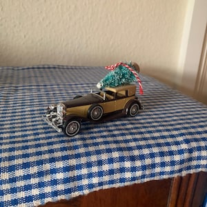 May include: A miniature brown and gold car with a small green Christmas tree on top. The car is on a blue and white checkered tablecloth.