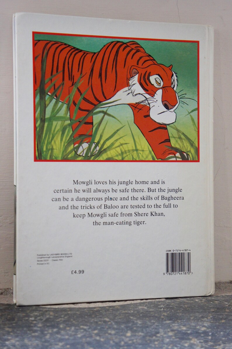 Puede incluir: La contraportada de un libro infantil sobre Mowgli de El Libro de la Selva. La portada presenta una ilustraci&oacute;n de un tigre caminando por la hierba alta y verde. El texto en la contraportada dice: "Mowgli ama su hogar en la selva y est&aacute; seguro de que siempre estar&aacute; a salvo all&iacute;. Pero la selva puede ser un lugar peligroso y las habilidades de Bagheera y los trucos de Baloo se ponen a prueba para mantener a Mowgli a salvo de Shere Khan, el tigre devorador de hombres."