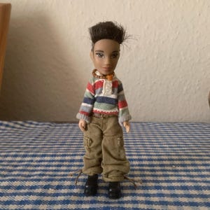 Könnte beinhalten: Eine Puppe mit dunkler Haut, bekleidet mit einem gestreiften Oberteil in Blau-, Rot- und Grautönen, kombiniert mit khakifarbenen Cargohosen und schwarzen Stiefeln. Die Puppe hat kurze, dunkle Haare und eine Perlenkette. Sie steht auf einer blau-weiß karierten Oberfläche.