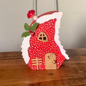 May include: A small red and white fairy house with a white snowy roof, a brown door, and a green holly sprig. The house is decorated with white polka dots.