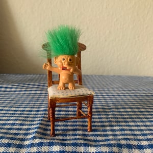 May include: A green-haired troll doll sitting on a wooden chair with a blue and white checkered pattern tablecloth underneath.