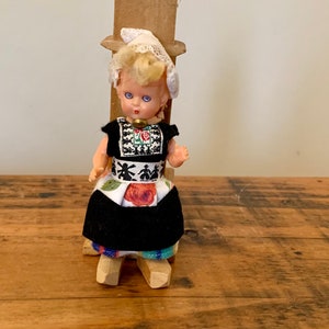 Könnte beinhalten: Eine Vintage-Puppe im niederländischen Stil, gekleidet in traditioneller Kleidung, steht auf Holzschuhen. Die Puppe hat blondes Haar, blaue Augen und trägt ein schwarzes Kleid mit floralen und gestickten Details. Eine weiße Spitzenkappe ziert ihren Kopf.