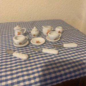 Peut inclure: Un service à thé miniature en porcelaine blanche avec un motif d'ours en peluche. L'ensemble comprend une théière, un sucrier, une crémière, des tasses, des soucoupes et des ustensiles de service. L'ensemble est présenté sur une surface à carreaux bleus et blancs.