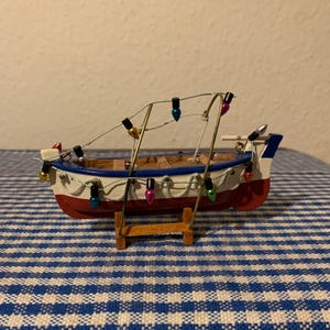 Puede incluir: Un pequeño modelo de barco de pesca de madera pintado de rojo, blanco y azul. El barco está decorado con una cadena de luces de colores y está sentado en un soporte de madera.