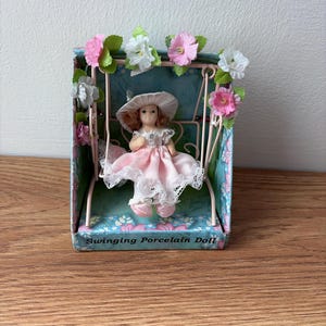 May include: A porcelain doll in a pink dress and hat sits on a swing inside a decorative box. The box is adorned with pink and white flowers and the text "Swinging Porcelain Doll".