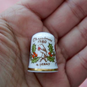 Puede incluir: Un dedal de cerámica blanca con borde dorado, con un diseño navideño. El dedal tiene un pájaro, acebo y el texto "Christmas 1980" y "M. Grant". Un artículo de colección.