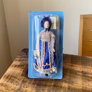 May include: A porcelain doll in a blue plastic package. The doll is dressed in a blue gown with gold trim, a matching hat, and carries a small purse. The doll has a fair complexion and dark hair. The packaging has a label.