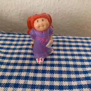 以下が含まれることがあります： 赤い三つ編みをしており、ピンクのリボンが付いた、小さな紫色のキャベツパッチキッズ人形の置物です。人形は「Love You Always」と書かれた小さな本を持っています。