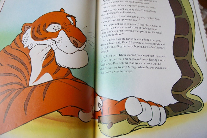 Puede incluir: Un primer plano de un tigre y una serpiente del libro "El libro de la selva". El tigre es naranja y negro con rayas blancas. La serpiente es verde y marr&oacute;n con un patr&oacute;n de escamas. El texto de la imagen dice: "&iquest;Con qui&eacute;n estabas hablando ah&iacute; arriba?" pregunt&oacute; Shere Khan. "Hablando conmigo mismo." respondi&oacute; Kaa. "Sabes que nunca te esconder&iacute;a nada, Shere Khan." dijo Kaa. Todo el tiempo, estaba desenrollando lentamente y con cuidado su cuerpo, con la esperanza de no molestar a Mowgli. Finalmente, Shere Khan pareci&oacute; convencido de que no hab&iacute;a nadie en el &aacute;rbol, y se fue, dejando a un Kaa muy asustado atr&aacute;s. Kaa estaba tan conmocionado que ni siquiera intent&oacute; detener a Mowgli cuando el ni&ntilde;o despert&oacute; y se desliz&oacute; por una enredadera para escapar.