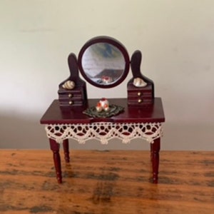 Puede incluir: Un tocador de madera en miniatura con un espejo redondo, dos cajones a cada lado y un borde de encaje. El tocador está pintado de color rojo oscuro y tiene un mantel de encaje blanco.