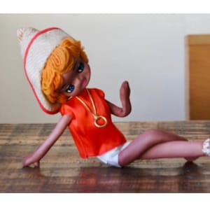 May include: A vintage doll with blonde hair, wearing a white and red hat, an orange shirt, and a gold necklace. The doll is lying on a wooden surface.