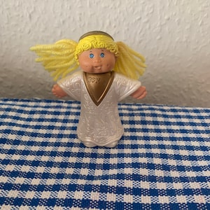Puede incluir: Una pequeña muñeca de ángel de plástico con cabello rubio, ojos azules y un vestido blanco con ribetes dorados. La muñeca tiene una cara de Cabbage Patch Kid y las letras "GPK" en su pecho.