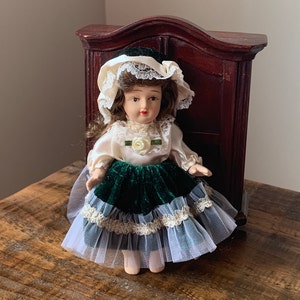 Może przedstawiać: Porcelanowa lalka z brązowymi włosami, ubrana w kremową sukienkę z zieloną aksamitną spódnicą i białą halką obszytą koronką. Lalka ma pasujący kapelusz i jest ustawiona przed ciemną drewnianą szafką.