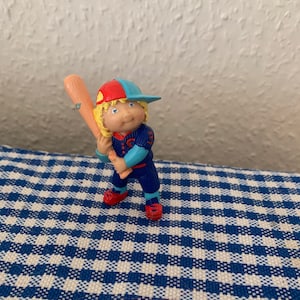 Può includere: Statuetta di un giocatore di baseball in uniforme blu, scarpe rosse e berretto rosso e blu, con una mazza di legno. La statuetta ha i capelli biondi ed è in posizione di battuta. Sfondo: muro bianco e tovaglia a scacchi blu e bianca.