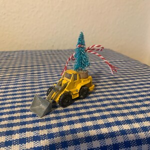 May include: A yellow toy construction vehicle with a blue and white Christmas tree on top. The vehicle has a silver scoop and is tied with a red and white string.