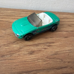 May include: A die-cast toy convertible car in a metallic teal finish with a white interior and a dark tinted windscreen. The vehicle features black wheels and blue and silver graphic decals along the side panels, showcasing a two-door sports car design.