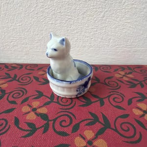 May include: A small, white porcelain cat figurine sits inside a matching bowl. The cat has blue accents on its ears and face. The bowl has a blue floral design around the rim. The figurine is on a red surface with a floral pattern.