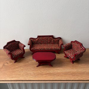 Op de afbeelding: Miniatuur poppenhuismeubelset. De set bevat een rode bank, twee fauteuils en een kleine ovale salontafel. De meubels zijn bekleed met een rood bloemenpatroon en hebben een donkerrood houten frame.