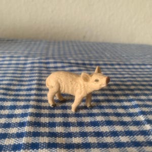 Op de afbeelding: Een kleine, lichtbruine varkensfiguur. Het varkentje staat op vier poten, met zijn snuit en oren duidelijk zichtbaar. De figuur staat op een blauw-wit geruite achtergrond.