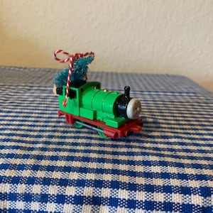 Könnte beinhalten: Ein grüner Spielzeug-Zugmotor mit einem kleinen Weihnachtsbaum oben drauf. Der Zug hat einen roten Schlusswagen und ein weißes Gesicht mit schwarzen Augen und einem Lächeln.