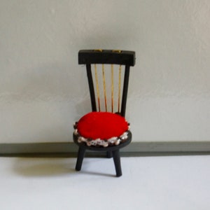 May include: A miniature black wooden chair with a red cushion and gold accents. The chair has a simple design with a rounded seat and four legs.