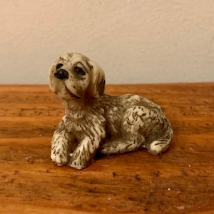 Peut inclure: Une petite figurine en céramique blanche d'un chien assis la tête levée. Le chien a une fourrure brune et des yeux noirs.