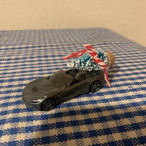 Op de afbeelding: Een grijze speelgoedauto met een kleine, met vlokken bedekte kerstboom die met rood en wit touw op het dak is vastgebonden.