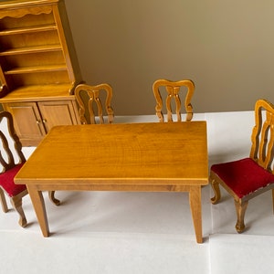 Puede incluir: Un juego de comedor de madera con una mesa rectangular y cuatro sillas. La mesa y las sillas son de color marrón claro, y las sillas tienen cojines rojos.