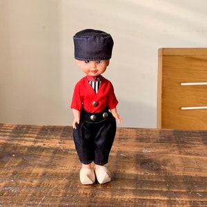 Könnte beinhalten: Eine kleine Puppe, die ein rotes Hemd mit weißen Streifen, eine schwarze Hose und einen blauen Hut trägt. Die Puppe trägt Holzschuhe und steht auf einem Holztisch.