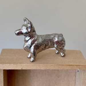 May include: A silver metal figurine of a corgi dog standing on a wooden shelf.