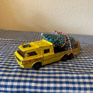 Puede incluir: Un camión de juguete amarillo vintage con un árbol de Navidad asegurado en la parte trasera con hilo rojo y blanco. El camión tiene ruedas negras y un diseño detallado. El árbol es verde con detalles blancos.