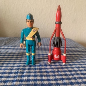 Puede incluir: Una figura de acción vintage de un hombre con uniforme turquesa con una faja amarilla y una gorra azul, junto a un cohete rojo con el número 3. Los juguetes están sobre una superficie a cuadros azules y blancos.