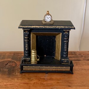 May include: A small, black fireplace with gold detailing, a miniature gold-coloured jug inside, and a tiny clock on top. The fireplace is rectangular with decorative columns and a detailed base. The clock has a white face and black hands.