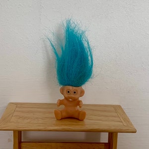 May include: A small plastic troll doll with blue hair sitting on a wooden table. The troll has a wide smile and large eyes.