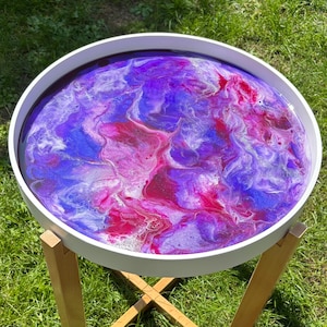 May include: A round white side table with a vibrant, swirling epoxy resin top in shades of purple, magenta, and blue. The table has a simple, natural wood base with an X-shaped support. The table is outdoors on a grassy surface.