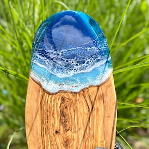 May include: An oval-shaped wooden serving board with a resin ocean design. The top half features a deep blue and white wave pattern, while the bottom half is natural wood grain. The board is ideal for serving appetizers or as a decorative piece.