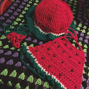 May include: A handmade crochet watermelon-themed set, including a hat and a triangle top. The hat is red with a green and white brim, and the top is a watermelon slice design. The set is displayed on a black, green, and purple crocheted blanket.