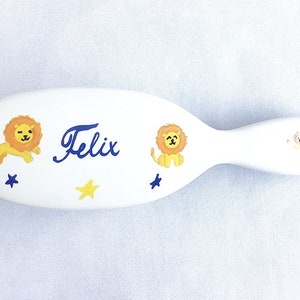 May include: A white baby hairbrush with a curved handle. The brush is decorated with yellow lions, blue stars, and a yellow star. The name "Felix" is written in blue script. A small lion is on the handle.