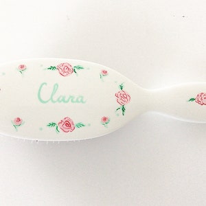 May include: White oval hairbrush decorated with pink roses and green leaves. The name "Clara" is written in light blue script. The brush has a handle and is designed for hair care.