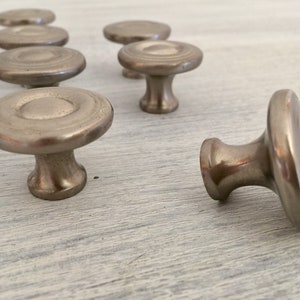 May include: A group of eight silver-toned cabinet knobs with a round, slightly raised center and a circular base. The knobs are arranged in a semi-circle on a light wood surface.