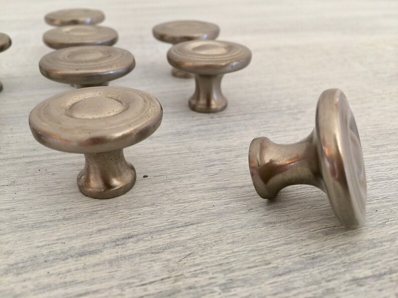 May include: A group of eight silver-toned cabinet knobs with a round, slightly raised center and a circular base. The knobs are arranged in a semi-circle on a light wood surface.