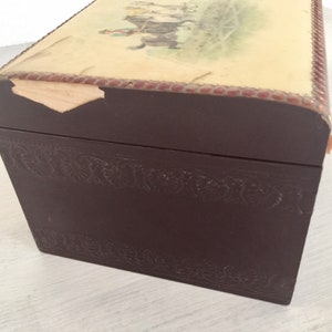 May include: A dark brown wooden box with a decorative embossed design around the edges. The top of the box is covered in a light beige paper with a faded image of a horse and rider. The paper is slightly torn on the top edge.