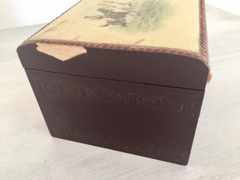 May include: A dark brown wooden box with a decorative embossed design around the edges. The top of the box is covered in a light beige paper with a faded image of a horse and rider. The paper is slightly torn on the top edge.
