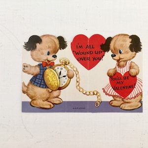 May include: A vintage Valentine's Day card featuring two cartoon dogs. One dog holds a pocket watch with a chain attached to a heart-shaped card that reads "I'm all wound up over you!" The other dog holds a heart-shaped card that reads "You'll be my Valentine!"
