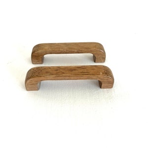 May include: Two wooden cabinet pulls with a rounded rectangular shape. The pulls are a light brown color and have a smooth finish.
