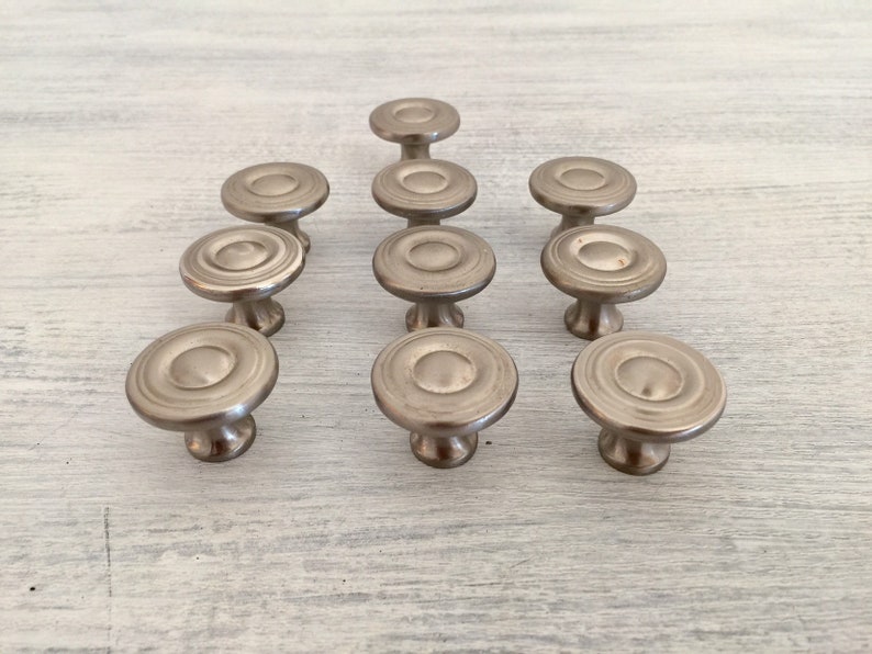 May include: Twelve silver cabinet knobs with a circular design and a raised center. The knobs have a satin finish and are arranged in a triangular pattern.