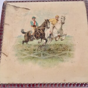 May include: A vintage wooden box with a red border and a printed image of two riders on horseback jumping over a fence. The riders are wearing jockey outfits and the horses are in mid-air.