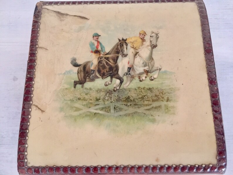 May include: A vintage wooden box with a red border and a printed image of two riders on horseback jumping over a fence. The riders are wearing jockey outfits and the horses are in mid-air.