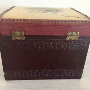 May include: A small wooden box with a burgundy lid and a floral design on the top. The box has brass hinges and a brown embossed design on the bottom.